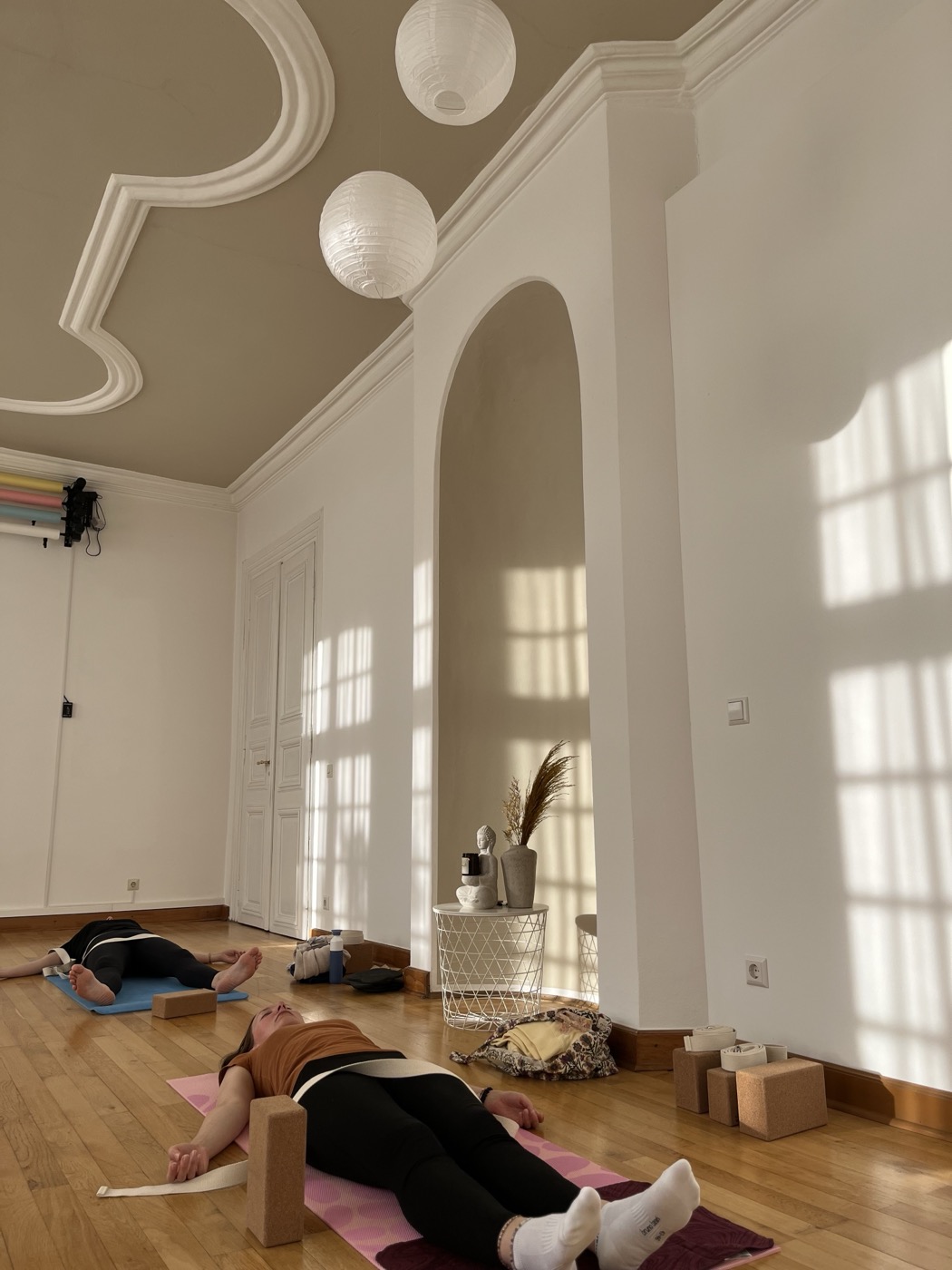 Zwei Teilnehmerinnen entspannt in Rückenlage mit Yoga-Blöcken, im lichtdurchfluteten Kursraum